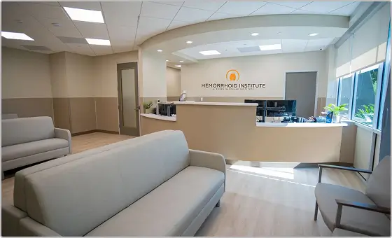 A modern medical office lobby with a light gray sofa, wood flooring, and a reception desk. The wall behind the desk displays the Hemorrhoid Institute logo and name. The space is well-lit with ceiling lights.